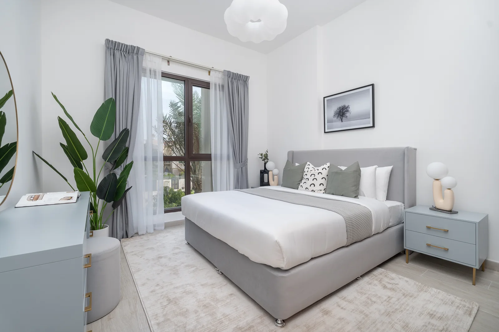 Bedroom with a grey upholstered bed, pale grey bedside cabinet and plants by the window.