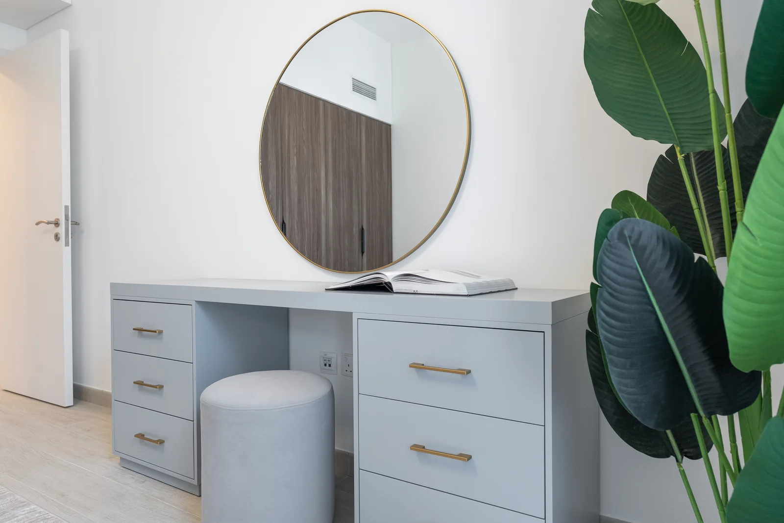 Pale grey dressing desk with gold-coloured handles, a round mirror and an upholstered stool.