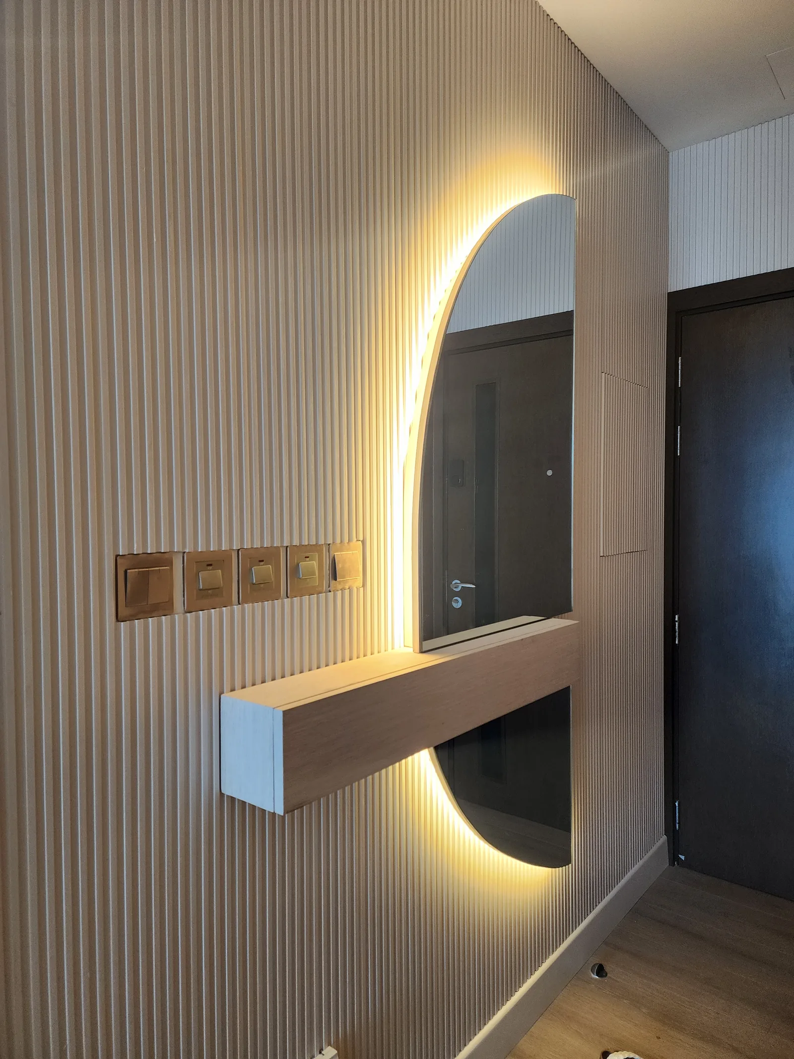 Oval mirror lit from behind and a floating shelf on vertically fluted panels.