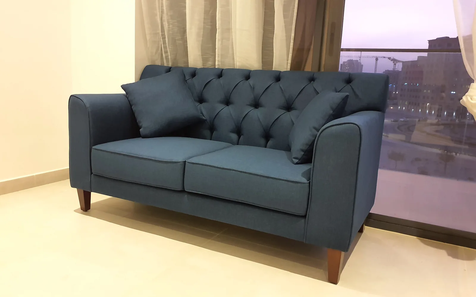 Blue two-seat sofa with a button-tufted back beside a window.