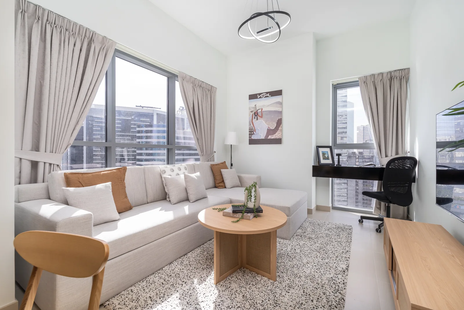 Pale living-room seating, round timber-toned table, curtains and a desk by the windows.