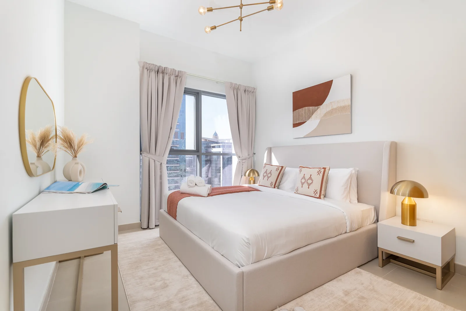 Bedroom with an upholstered winged bed, white bedside table, gold-toned lamps and a white desk by the window.