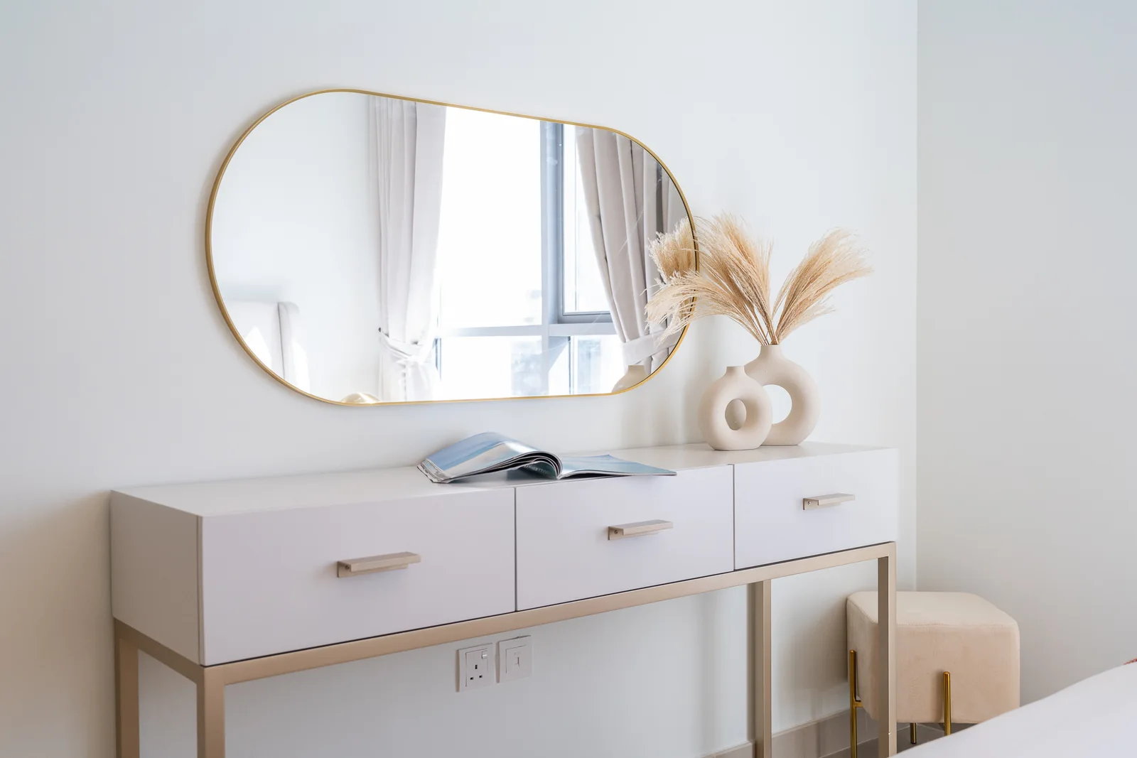 White three-drawer dressing table on a champagne-toned metal frame with a gold-toned capsule mirror and cream stool.