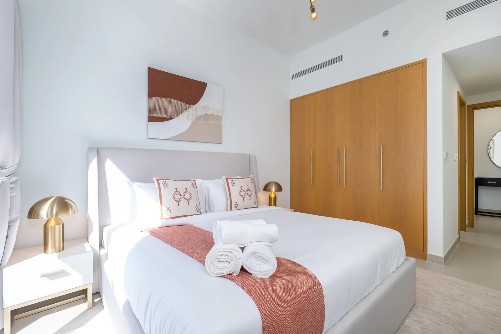 Upholstered bed with a terracotta throw beside a four-door timber-toned wardrobe.