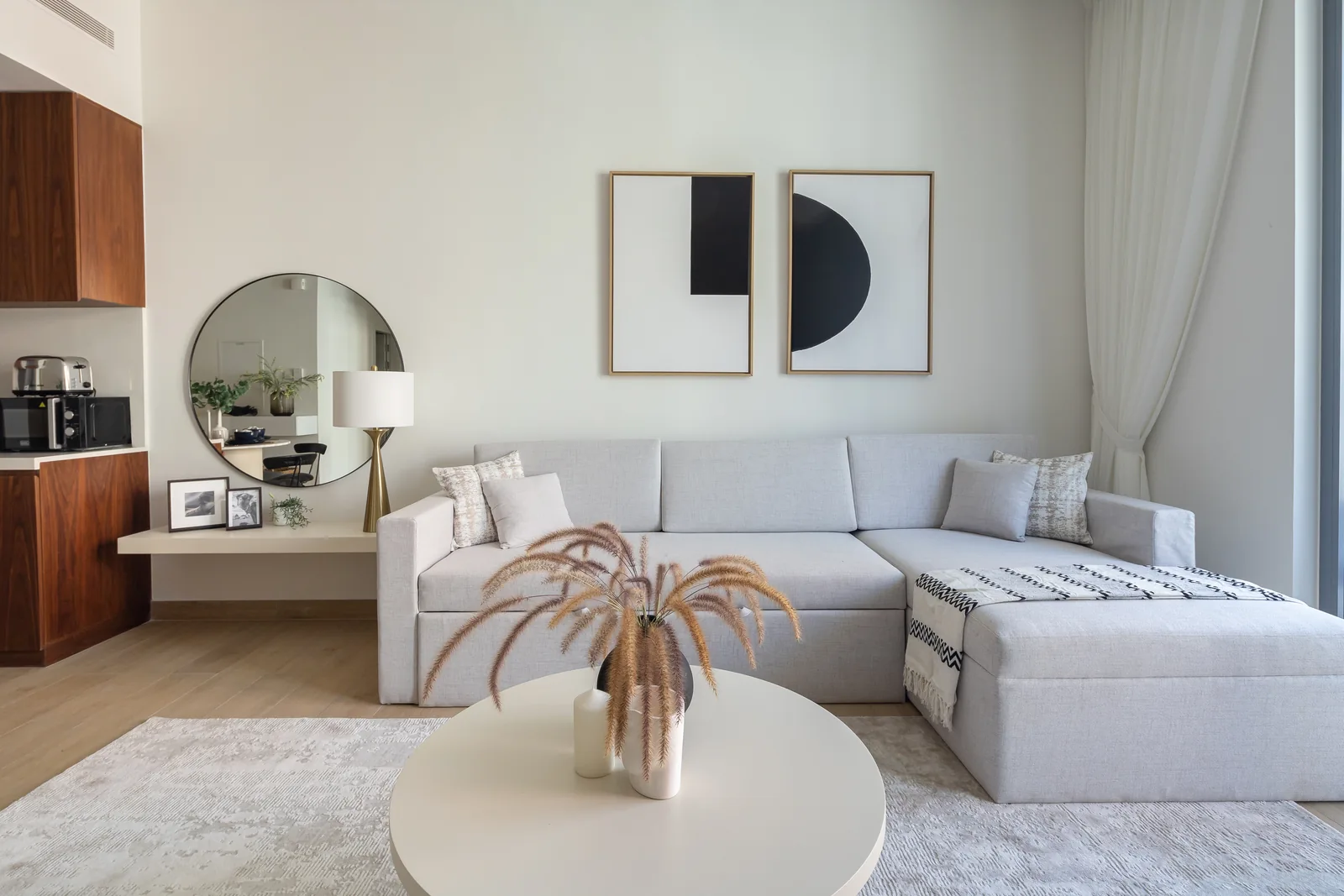 Pale sectional sofa, round table and circular mirror beside timber-toned cabinetry.