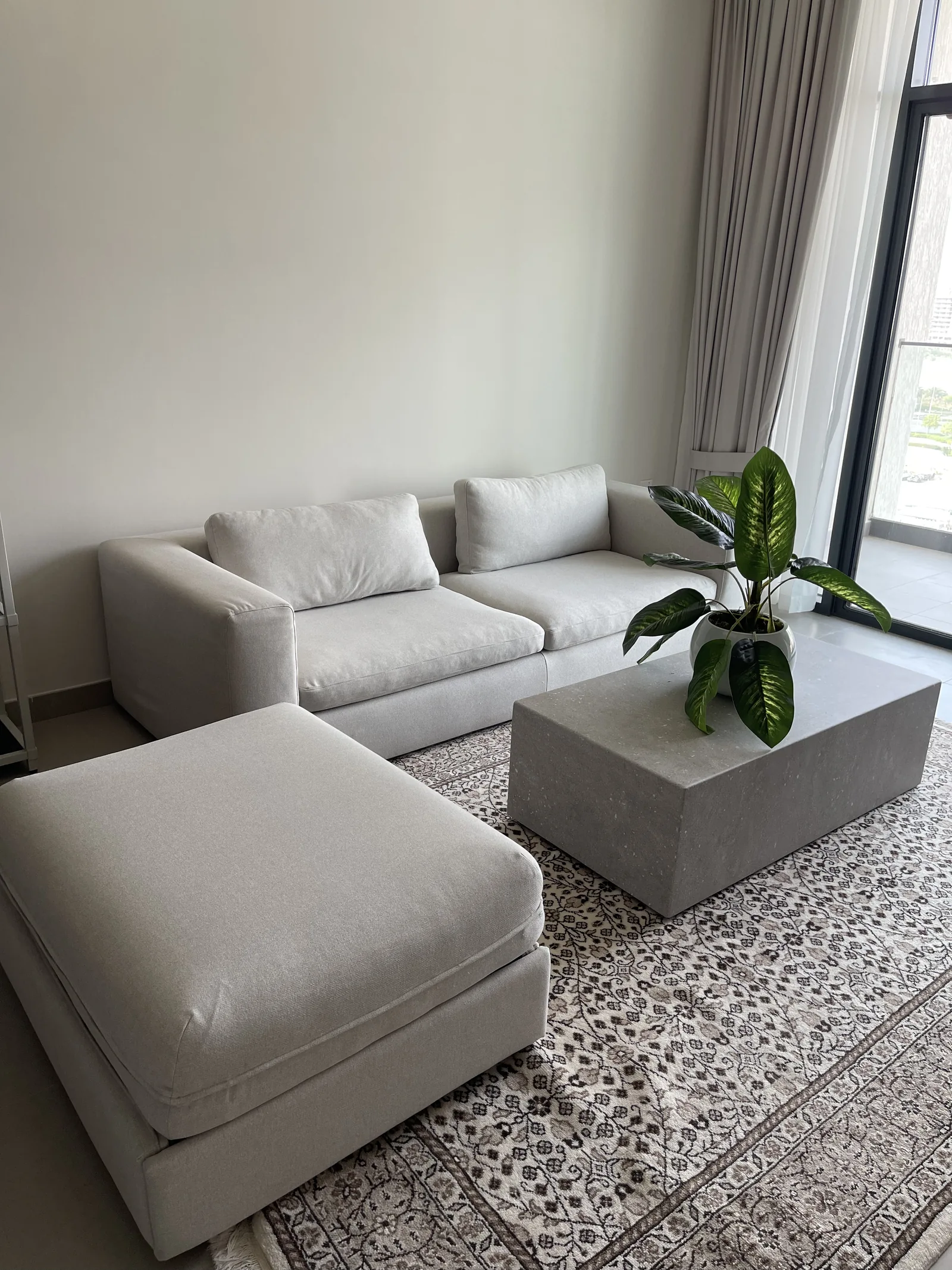Pale sectional and ottoman around a grey block coffee table on a patterned rug.