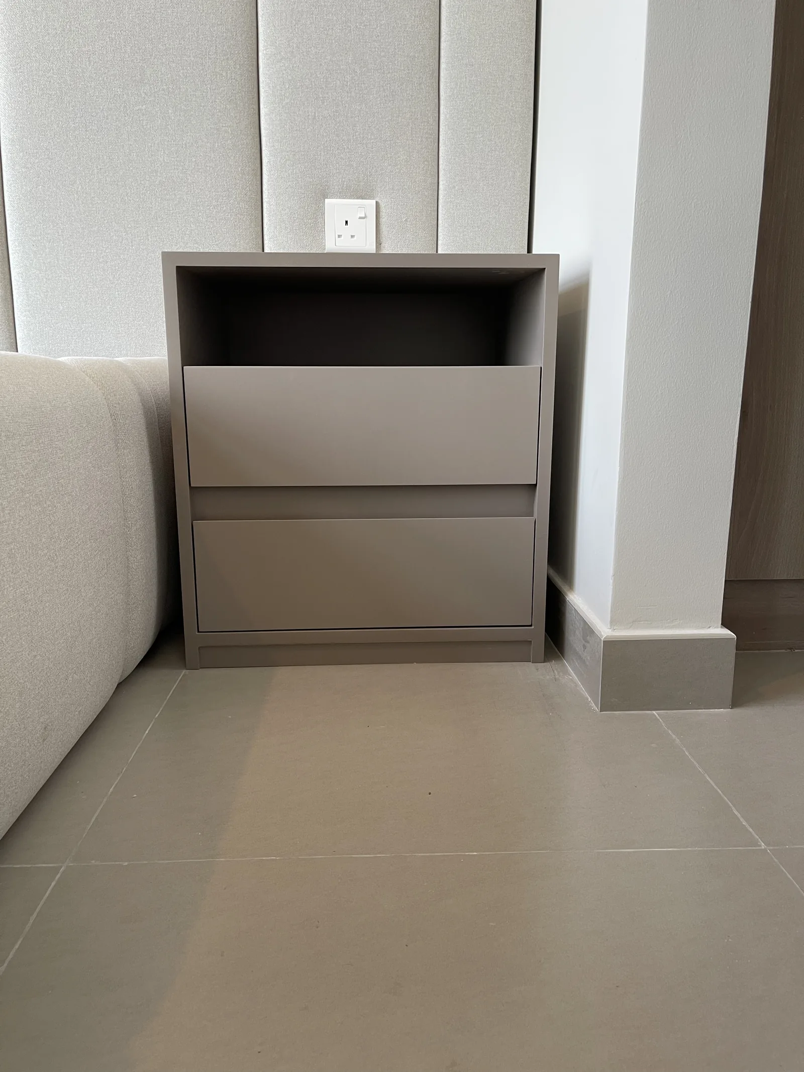 Pale grey bedside cabinet with an open shelf and drawers beside an upholstered wall panel.