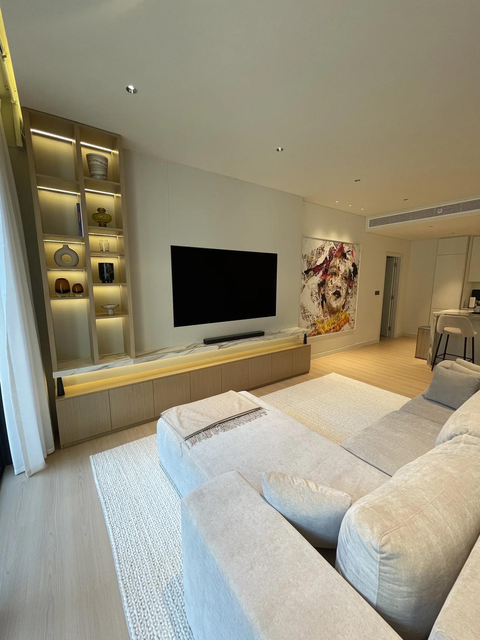 Illuminated display shelves beside a low timber-toned media cabinet, with a pale sectional in front.