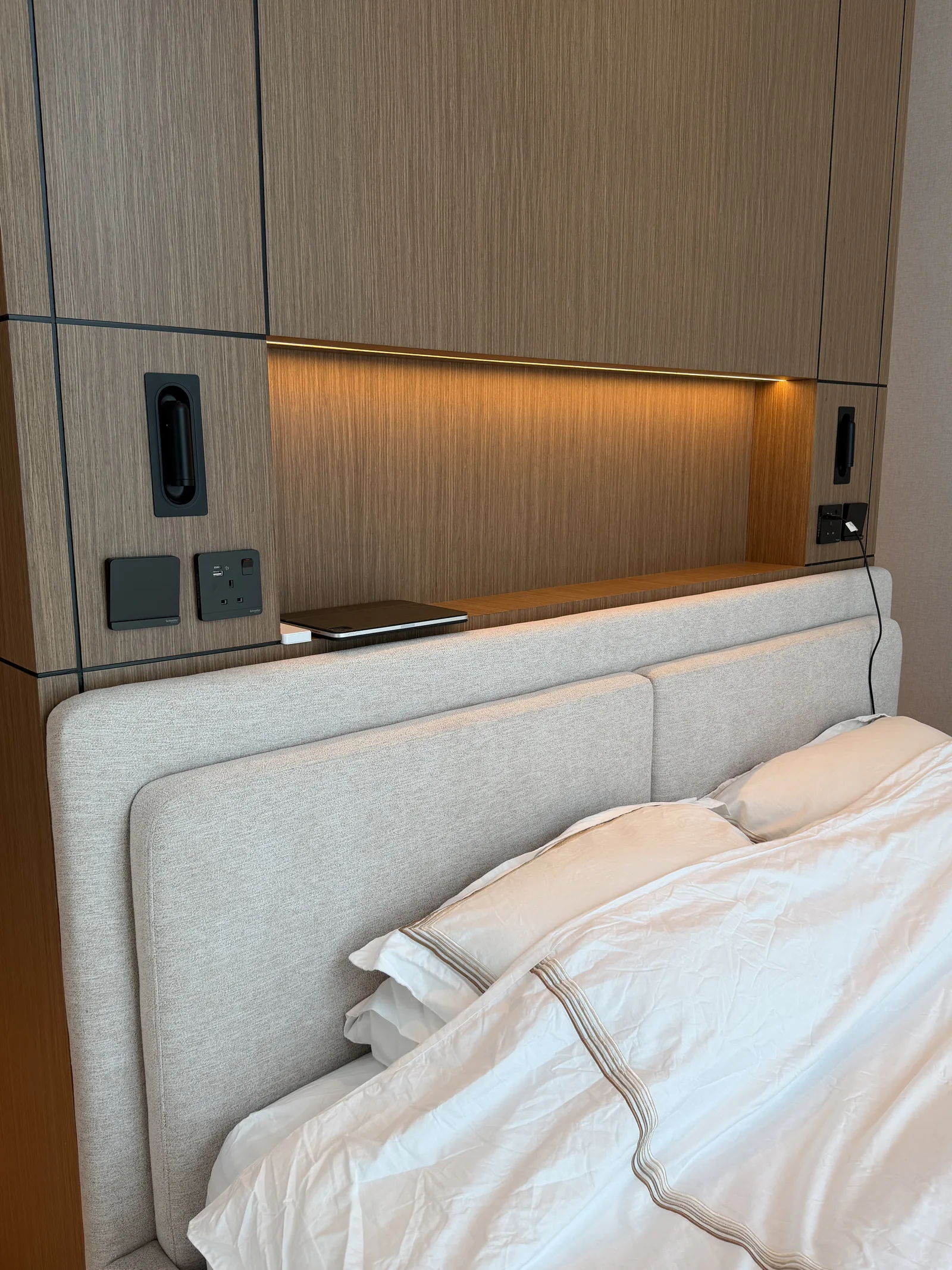 Pale upholstered headboard beneath timber-toned wall panels and a lit shelf.