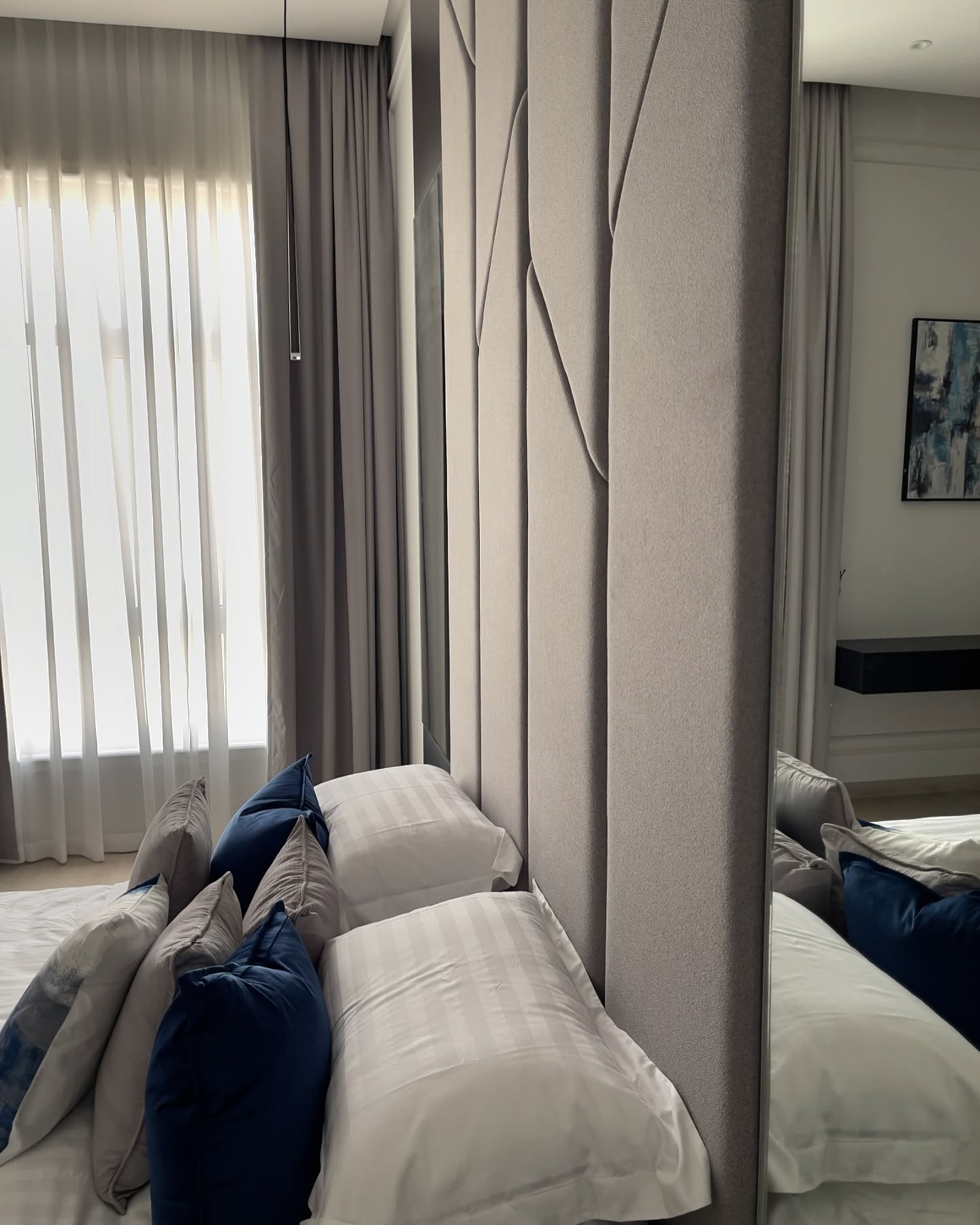 Close-up of a geometric upholstered headboard beside a full-height mirror and sheer curtains.